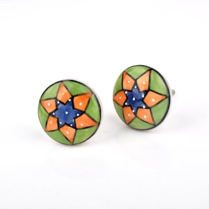 May include: Two decorative ceramic cabinet knobs. Each knob is round with a green background and a starburst design in orange and blue. The starburst has white dot accents. The knobs have silver metal hardware.