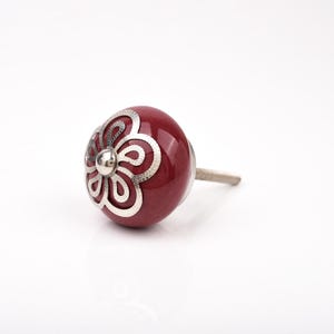May include: A burgundy cabinet knob with a spherical shape. It has a silver floral pattern on the front and a silver stem. The knob is designed for use on cabinets, drawers, or furniture. The knob's dimensions are in centimetres.