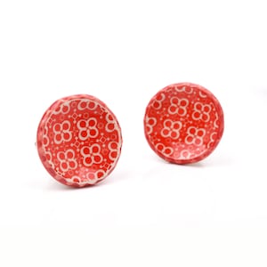 May include: Two round, concave ceramic drawer knobs featuring a vibrant red background with a repeating white geometric pattern of four-petaled flower shapes and small diamond accents. The edges of the knobs have a slightly irregular, textured finish.