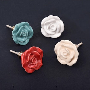 May include: Four drawer knobs shaped like blooming roses in teal, white, red, and cream colors. Each knob features a sculpted floral design with layered petals and a metallic screw post extending from the back for attachment to furniture.
