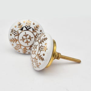 Peut inclure: Deux boutons de meuble en céramique blanche avec des motifs floraux dorés. Les boutons sont ronds, avec une base en laiton et une tige filetée pour l'installation. Le design présente des détails dorés complexes.