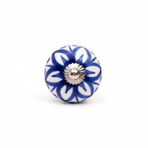 May include: A round ceramic cabinet knob featuring a white base with a hand-painted blue floral pattern. The center of the knob has a silver-toned metal hardware piece with a scalloped edge and a smooth, rounded top surface.