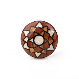 Può includere: Pomello per armadio rotondo in ceramica con design geometrico. Il pomello presenta uno sfondo marrone con un motivo di forme bianche, beige e marroni. Un cerchio bianco è al centro, delineato in nero.