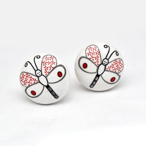 Puede incluir: Dos pomos de armario de cerámica blanca con diseño de mariposa. La mariposa tiene alas estampadas en rojo y blanco, cuerpo negro y contornos grises. Los pomos son redondos y tienen un acabado brillante.