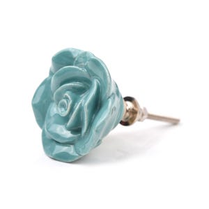 May include: A teal-colored ceramic cabinet knob shaped like a blooming rose. The floral-shaped handle features a glossy finish and is attached to a silver-toned metal bolt with a washer and nut for installation on furniture or cabinetry doors.