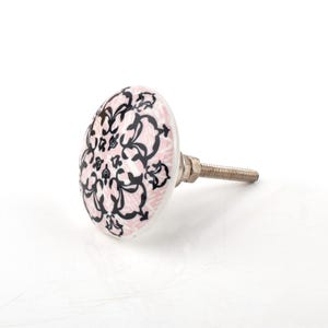 May include: A decorative cabinet knob with a pink and black floral design. The knob is round with a white edge and a metal screw for installation. The design features a repeating pattern of black floral elements on a pink background.