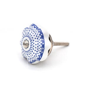 May include: White ceramic cabinet knob with a blue geometric pattern. The knob has a flower-like shape with a silver metal screw. The blue pattern includes small crosses and dots. This decorative knob is ideal for furniture.