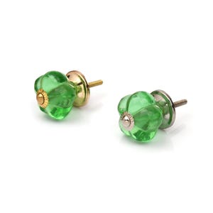 Puede incluir: Dos pomos de cristal verde para armarios con forma acanalada similar a una calabaza. Cada pomo presenta una tapa central de metal dorado con borde estriado y un tornillo metálico roscado que sobresale por detrás para su instalación.