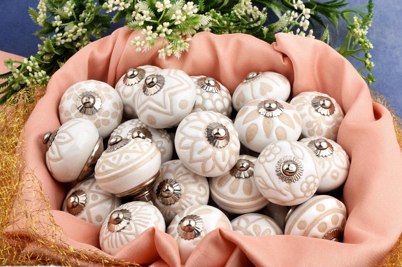 collection of white ceramic drawer knobs with intricate floral patterns and silver accents. The knobs are arranged in a basket lined with peach-colored fabric.