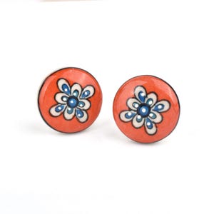 May include: Two round orange cabinet knobs with a floral design. The flower has white and blue petals with a dark blue center. The knobs are likely made of ceramic or a similar material.