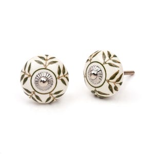 May include: Two round cabinet knobs with a cream-coloured base and a dark green leaf pattern. Each knob features a central silver-toned metal cap with a fluted edge and a small screw protruding from the back of one of the knobs for installation.