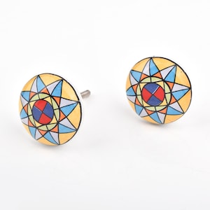 Könnte beinhalten: Zwei dekorative Schrankknöpfe mit einem farbenfrohen geometrischen Design. Die Knöpfe haben einen gelben Hintergrund mit einem Sternmuster in Blau, Orange, Rot und Grün. Jeder Knopf hat einen weißen Sockel und eine silberne Schraube.