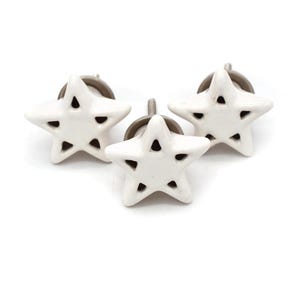 Puede incluir: Tres pomos de cajón de cerámica blanca en forma de estrella con un diseño recortado. Los pomos tienen un tornillo de metal plateado.