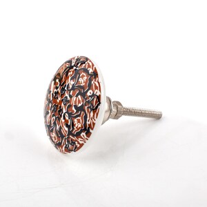 May include: A ceramic cabinet knob featuring a brown, black, and white floral design. The knob has a metal screw for attaching to furniture. The pattern is a repeating design of stylized leaves and swirls.