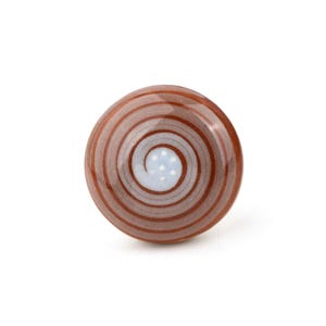 May include: A round ceramic cabinet knob with a spiral design. The knob has concentric brown and white stripes, with a light blue centre containing white dots. It is a decorative element for furniture.