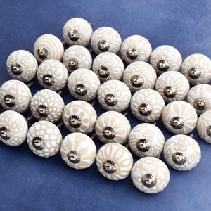 collection of white ceramic drawer knobs with intricate floral patterns and silver accents. The knobs are arranged in a basket lined with peach-colored fabric.