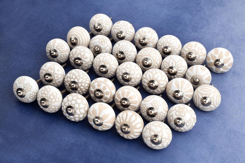 collection of white ceramic drawer knobs with intricate floral patterns and silver accents. The knobs are arranged in a basket lined with peach-colored fabric.