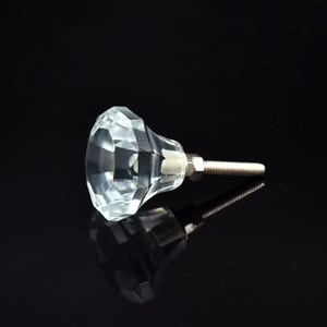 May include: A clear, faceted crystal cabinet knob with a silver screw and nut. The knob has a diamond-like shape and reflects light. The hardware is designed for easy installation on drawers or cabinets. The knob's dimensions are in centimetres.