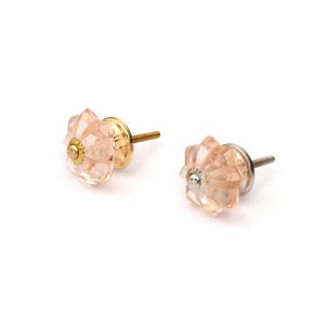 May include: Two faceted, light pink glass cabinet knobs with a star-like shape. Each knob features a central metallic cap and a threaded metal screw extending from the back. One knob has a gold-toned base, while the other has a silver-toned base.