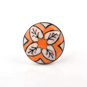 Può includere: Un pomello rotondo in ceramica per mobili con un vivace motivo floreale arancione e bianco. Il design presenta dettagli neri simili a rami e piccoli punti rossi disposti all'interno di petali bianchi su uno sfondo arancione brillante.