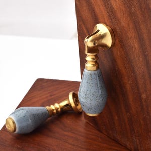 May include: Two cabinet pulls with a brass finish and speckled blue ceramic handles. One pull is mounted on a dark wood surface, while the other lies horizontally. The pulls have a teardrop shape.