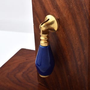May include: A cabinet pull with a brass fitting and a deep blue, teardrop-shaped ceramic handle. The pull is mounted on a dark wood surface, highlighting the contrast. The hardware is designed for furniture, with a focus on the handle's design.