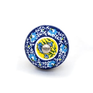 Può includere: Un pomello per armadio rotondo in ceramica con un motivo floreale. Il pomello presenta uno sfondo blu scuro con motivi floreali bianchi e blu chiaro. Il centro ha un cerchio giallo con fiori blu e un centro in metallo argentato. Il pomello misura circa 3,8 cm di diametro.