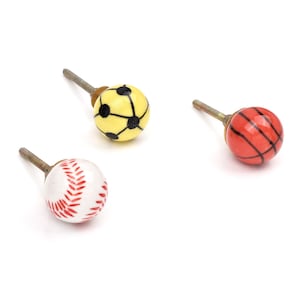 Op de afbeelding: Drie bolvormige kastknoppen met sportbalontwerpen. Eén knop is wit met rode honkbalstiksels, één is geel met zwarte voetbalpatronen en één is oranje met zwarte basketbal-lijnen, elk bevestigd aan een metalen bout.
