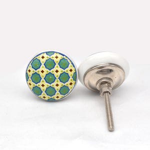 May include: Two ceramic cabinet knobs with a white base and a decorative patterned top. The pattern features a teal and yellow geometric design. One knob is shown with the screw attached.