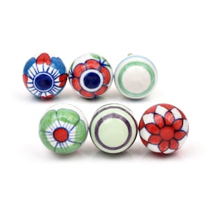 Op de afbeelding: Een set van zes ronde keramische knoppen met diverse handgeschilderde ontwerpen. De patronen omvatten blauwe en rode bloemmotieven, groene en paarse concentrische cirkels, een groene bloem met een rood hart en een rood-wit geblokt bloemmotief.