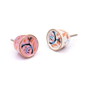 Op de afbeelding: Twee keramische kastknoppen met een conische vorm en een metalen schroef. De ene knop heeft een roze achtergrond en de andere een witte. Beide zijn voorzien van een geschilderd ontwerp van blauwe takken met oranje bladeren en kleine roze bloemen.