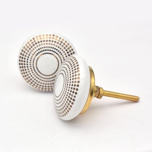 Può includere: Due pomelli per mobili in ceramica bianca con un motivo a pois dorati disposti in cerchi concentrici. I pomelli hanno una base in ottone e una vite filettata per l'installazione.