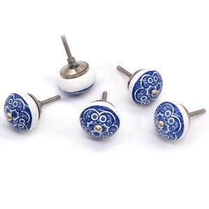 Könnte beinhalten: Fünf weiße und blaue Keramikschrankknöpfe mit floralem Design. Jeder Knopf hat eine silberfarbene Schraube und eine dekorative silberfarbene Mitte. Die Knöpfe sind auf weißem Hintergrund angeordnet.