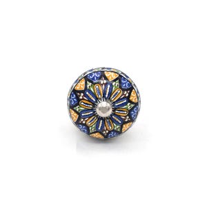 Könnte beinhalten: Ein dekorativer Schrankknopf mit floralem Design. Der Knopf hat einen silbernen Sockel und eine runde, gemusterte Oberseite mit blauen, gelben, grünen und orangen Farben. Das Design ist symmetrisch und detailliert.