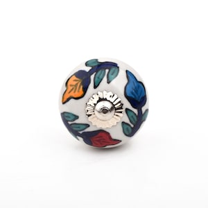 May include: A white ceramic cabinet knob with a floral design. The knob features orange, blue, and red leaves with dark green stems. A silver-coloured metal base is attached to the knob.
