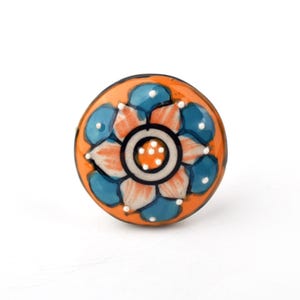 May include: A round ceramic cabinet knob featuring a vibrant orange background with a central floral pattern. The design includes alternating blue and white petals, accented with small white dots, and a dark circular ring around the centre.