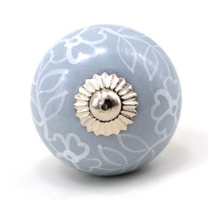 Könnte beinhalten: Hellblauer Keramik-Schrankknopf mit weißem Blumenmuster. Der Knopf hat einen silberfarbenen Metallsockel und eine dekorative, geriffelte Mitte. Diese dekorative Hardware ist ideal, um Möbeln einen Hauch von Eleganz zu verleihen.