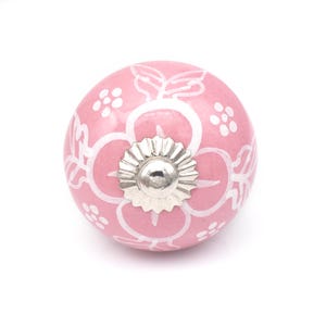 Puede incluir: Un pomo de armario de cerámica rosa con un diseño floral blanco. El pomo tiene una base de metal plateado y una forma redondeada. El patrón floral incluye hojas estilizadas y pequeños grupos de flores.