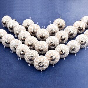 collection of white ceramic drawer knobs with intricate floral patterns and silver accents. The knobs are arranged in a basket lined with peach-colored fabric.