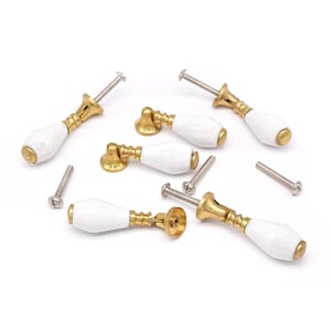May include: Several white and gold cabinet pulls with screws are scattered on a white surface. The pulls have a white, oval-shaped handle and a gold-colored base. The screws are silver.