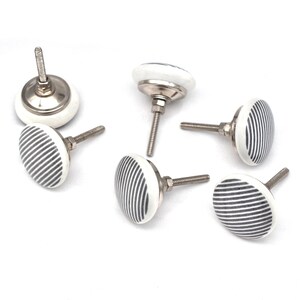 Op de afbeelding: Een set van zes witte keramische kastknoppen met een zilverkleurige metalen basis en schroef. Elke knop heeft een patroon van grijze verticale strepen. Ontworpen voor meubels en woondecoratie.