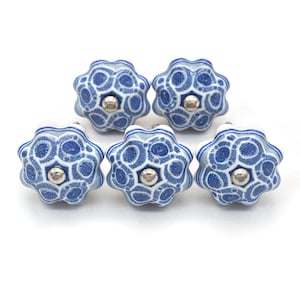 Op de afbeelding: Vijf blauwe en witte keramische kastknoppen met een bloemmotief. Elke knop heeft een zilverkleurig midden en een herhalend patroon van blauwe vormen op een witte achtergrond. Deze decoratieve knoppen zijn ideaal voor meubels.