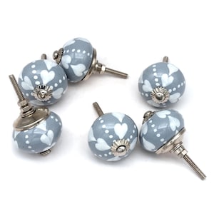May include: Six grey ceramic cabinet knobs with white heart designs. Each knob has a silver screw.
