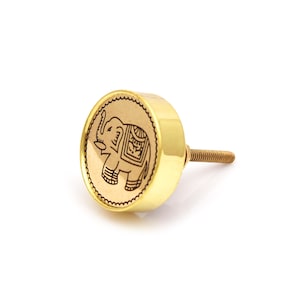 Peut inclure: Bouton de meuble doré avec un motif d'éléphant en relief. Le bouton a une face ronde avec un bord festonné et une tige filetée pour la fixation. L'éléphant est représenté avec des motifs détaillés et une trompe levée.