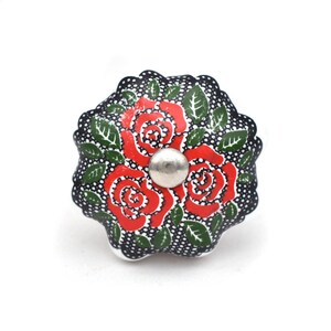 Peut inclure: Bouton de meuble décoratif en céramique avec un motif floral. Le bouton a un fond noir avec des pois blancs, des roses rouges, des feuilles vertes et un centre en métal argenté. Ce matériel ajoute une touche d'élégance.