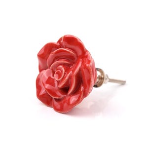 May include: A bright red ceramic drawer knob shaped like a blooming rose flower. The floral piece features layered, glossy petals and is attached to a silver-toned metal bolt with a nut, designed for installation on furniture or cabinetry hardware.