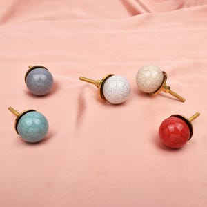May include: Five spherical cabinet knobs with a crackled glaze finish in blue, white, cream, and red. Each knob features a brass-toned metal base and a threaded screw attachment, showcasing a distinct network of fine lines across the ceramic surface.