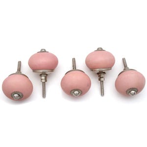 May include: Five pink ceramic cabinet knobs with silver-toned hardware. The knobs have a rounded shape and are arranged against a white background. These decorative knobs are suitable for furniture and home decor projects.