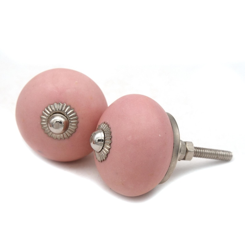 May include: Two pink ceramic cabinet knobs with silver-colored hardware. The knobs have a round shape with a decorative silver rosette around the screw. The knobs are set against a white background.
