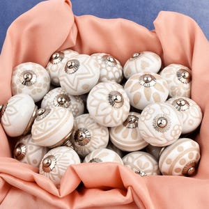 collection of white ceramic drawer knobs with intricate floral patterns and silver accents. The knobs are arranged in a basket lined with peach-colored fabric.